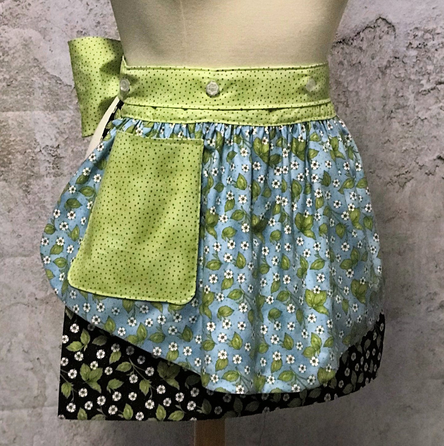 Ida’s Diva Apron is a patent pending convertible kitchen apron that can be worn 8 different ways by using the buttons on the waistband to flip, add or subtract both the apron front and back. Featuring generously sized pockets, 100% high quality cotton fabric, and long waist ties to make this apron well suited for most figures. With tiny flowers of lavenders and pinks and strawberry blossoms highlighted by a waistband of soft green teensy dots, this apron is a happy helper in any setting.
