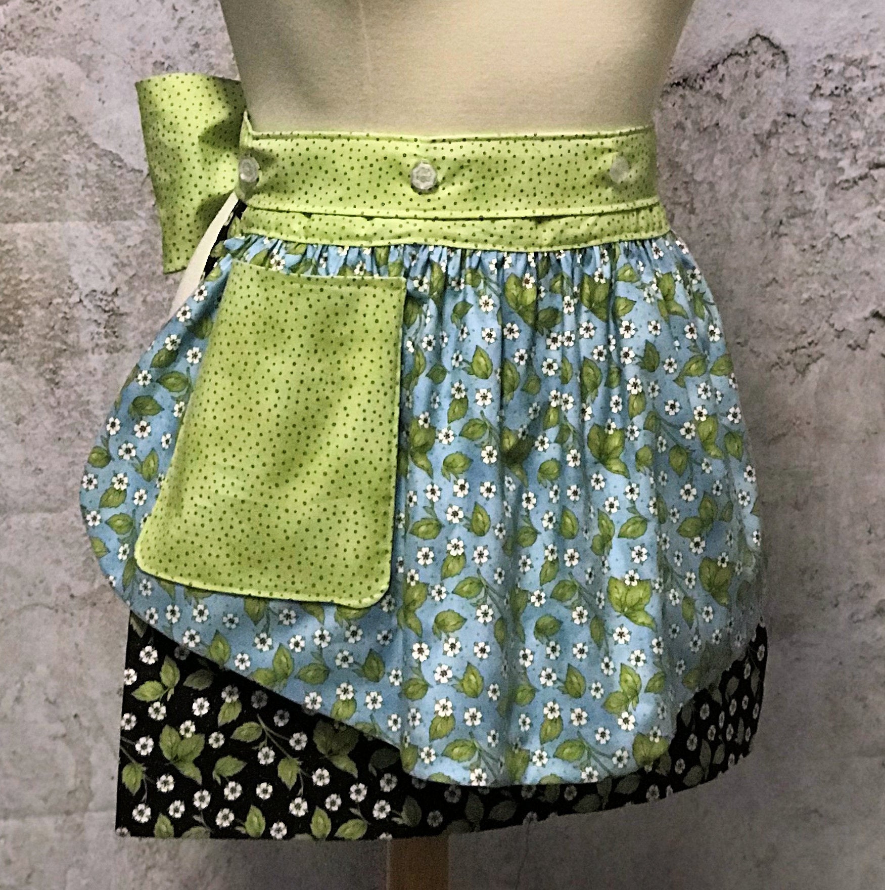 Ida’s Diva Apron is a patent pending convertible kitchen apron that can be worn 8 different ways by using the buttons on the waistband to flip, add or subtract both the apron front and back. Featuring generously sized pockets, 100% high quality cotton fabric, and long waist ties to make this apron well suited for most figures. With tiny flowers of lavenders and pinks and strawberry blossoms highlighted by a waistband of soft green teensy dots, this apron is a happy helper in any setting.